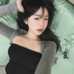 Malaysia Escort Girl Young woman with dark hair lying on a bed, wearing a black tube top and gray cardigan. Sex Service Kuala Lumpur