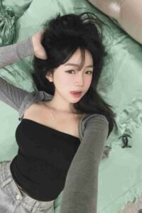 Malaysia Escort Girl Young woman with dark hair lying on a bed, wearing a black tube top and gray cardigan. Sex Service Kuala Lumpur