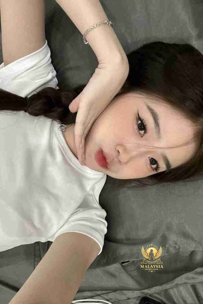 Asian woman with braid posing on gray bedding. Light skin, dark hair, wearing white t-shirt and bracelet.