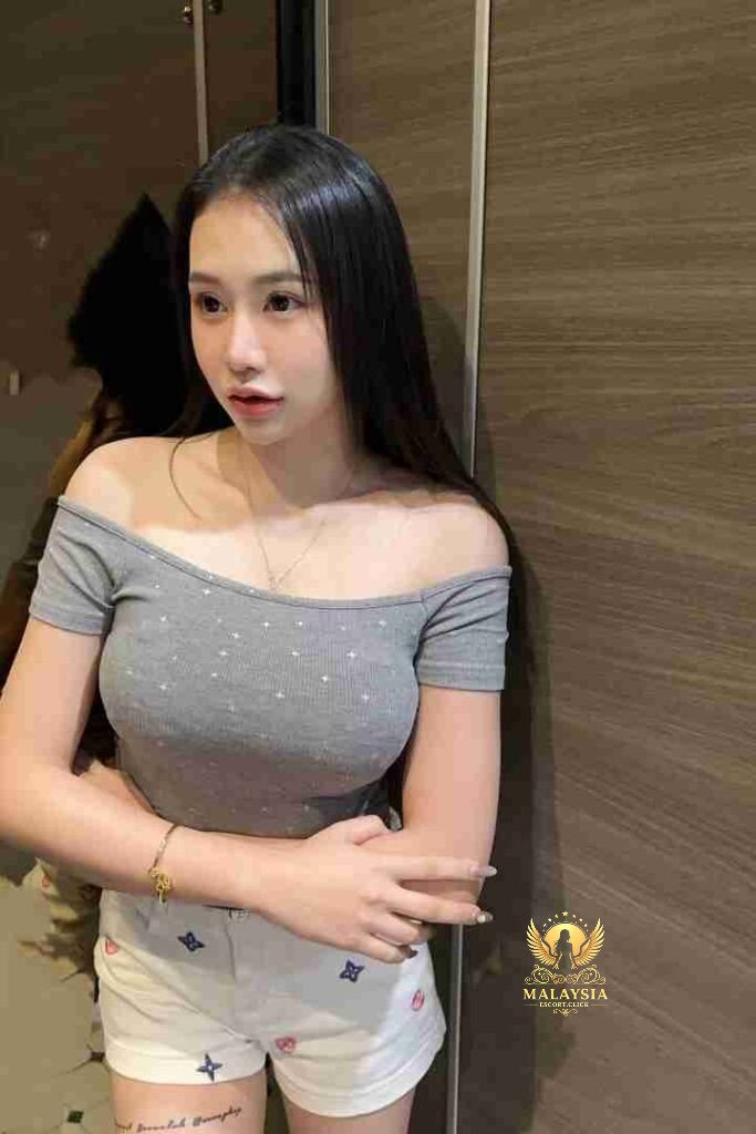 Attractive Asian woman in off-shoulder top and shorts, gold necklace and bracelet, posing indoors.