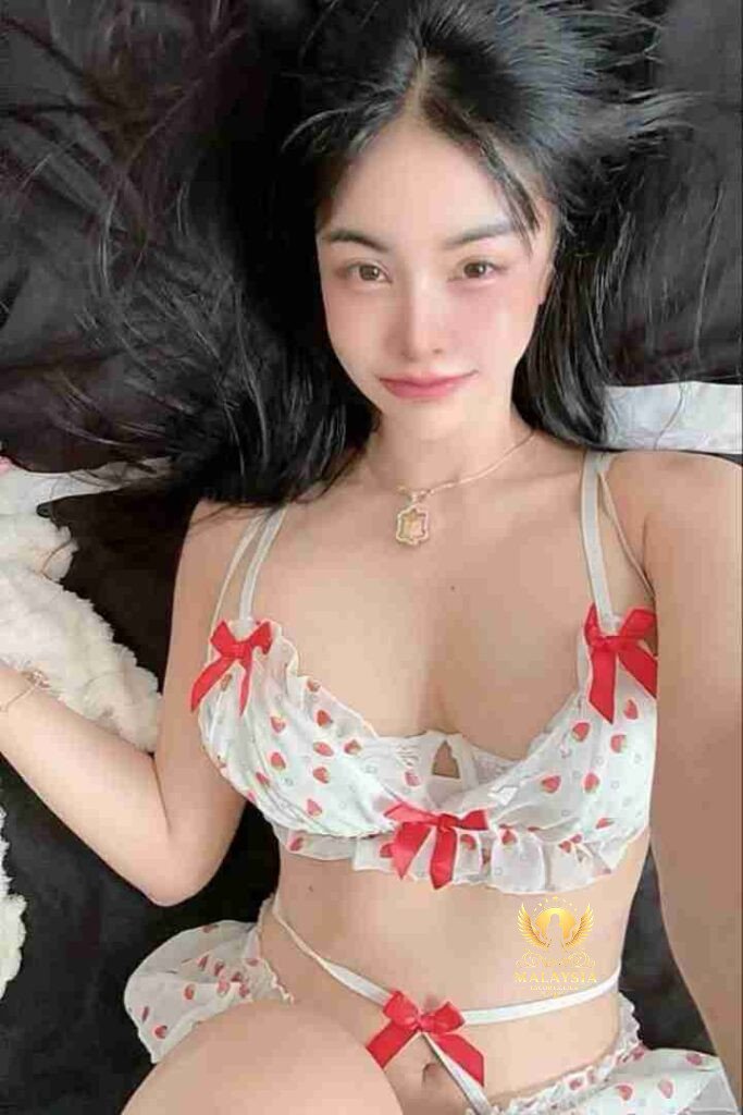 Asian woman in strawberry lingerie lying on bed, smiling at camera. Red bows, white fabric, black hair.