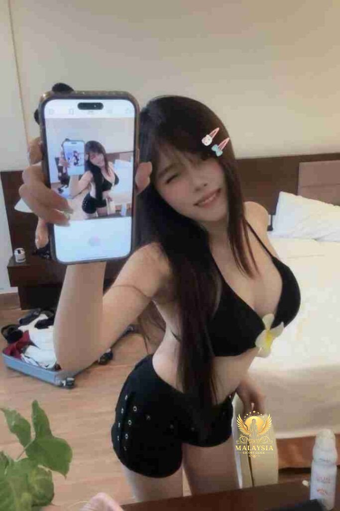 Woman in black bikini and shorts taking a selfie with her phone in a hotel room.