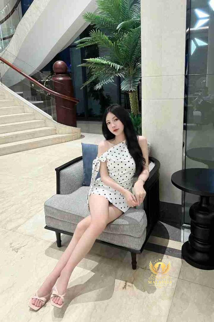Woman in polka dot dress sitting in chair, elegant hotel lobby with stairs and palm tree.